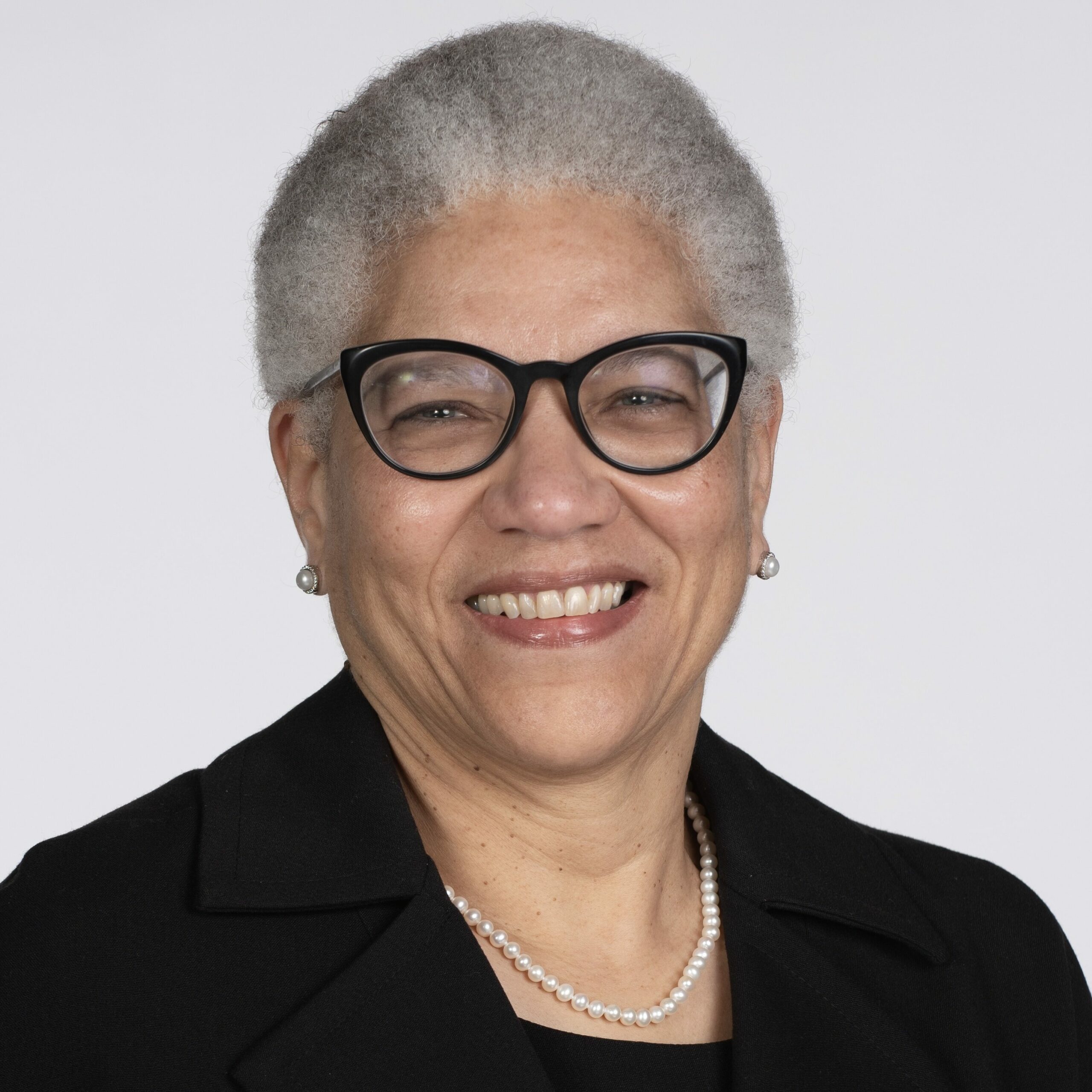 Portrait of a Black woman with short grey hair, wearing glasses and a pearl necklace, smiling at the camera