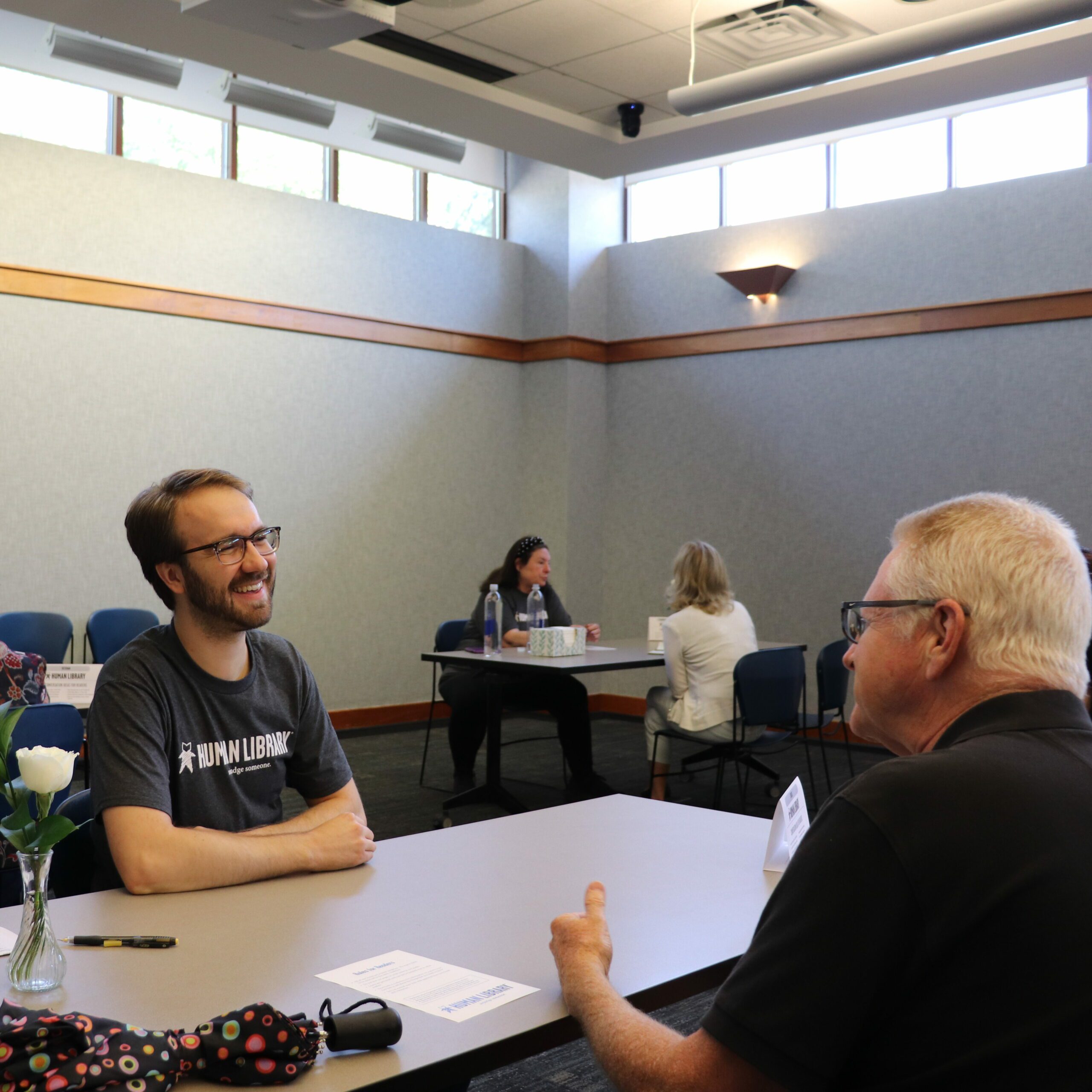 A man wearing a Human Library shirt sits at a table in the library's Meeting Room, smiling at another man, who sits across the table from him