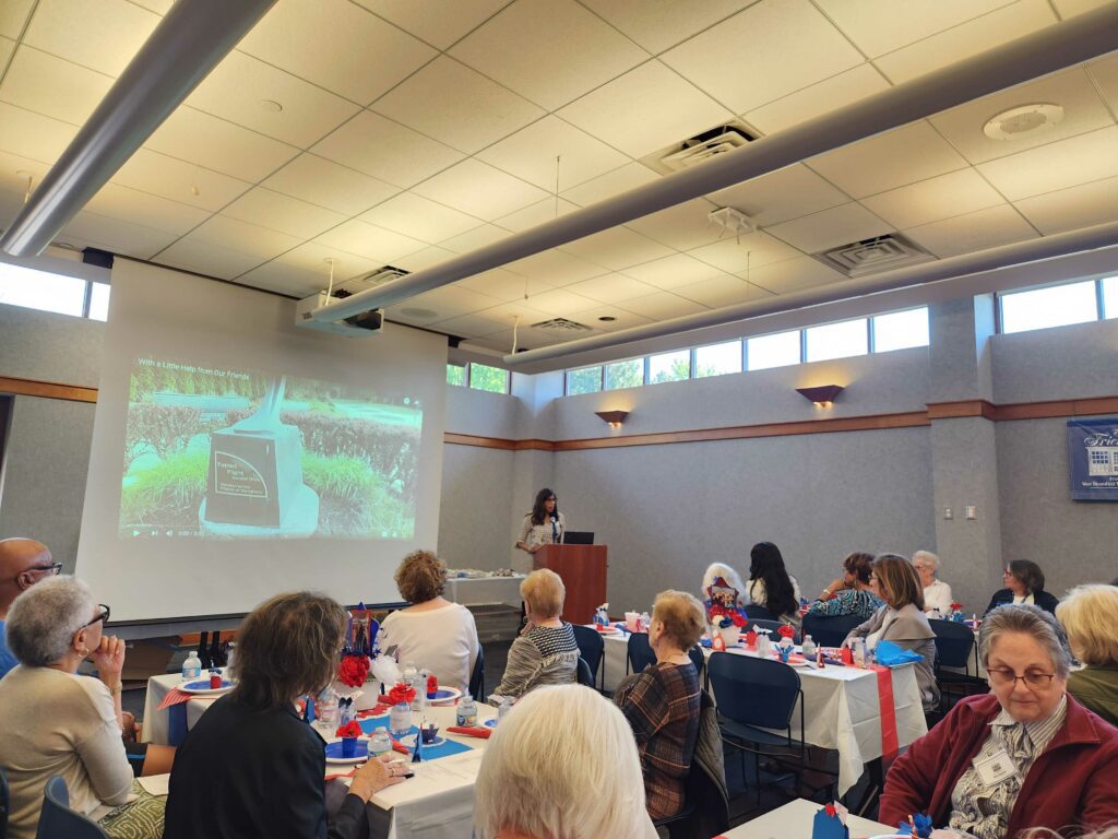 The Friends of the Library luncheon in the Main Library Meeting Room. President Johnnie Roquemore stands at the podium next to a screen. Tables are decorated in festive red, white, and blue colors. 