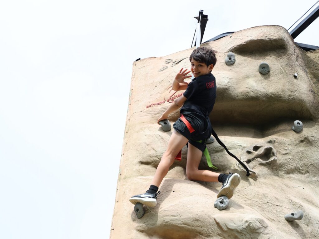 kid with short hair waving from the top of a rock climbing wall